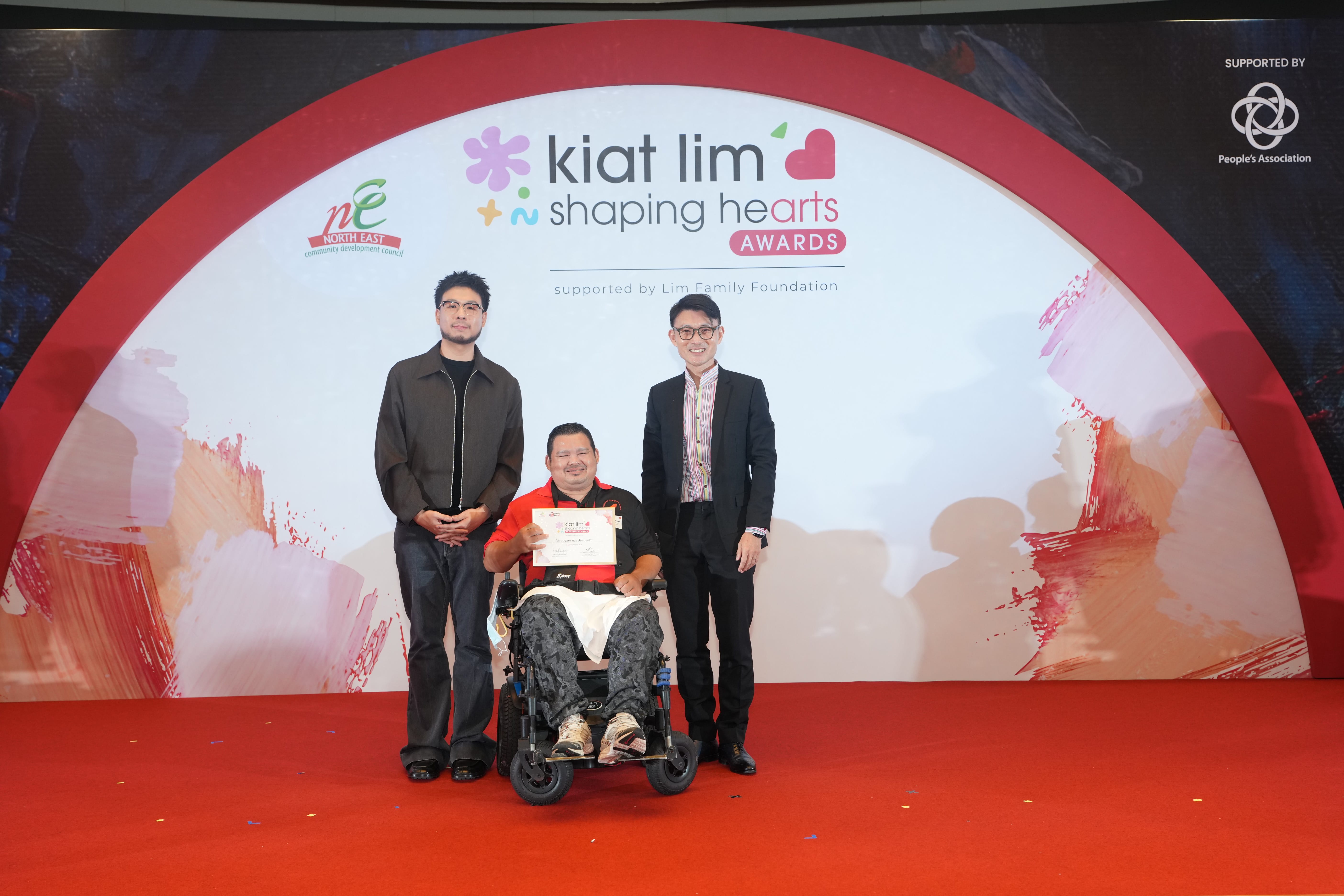 Award recipient standing on stage with the presenter, holding their certificate and posing for a group photo during the award ceremony, with a backdrop reading ‘Kiat Lim-Shaping Hearts Awards'.